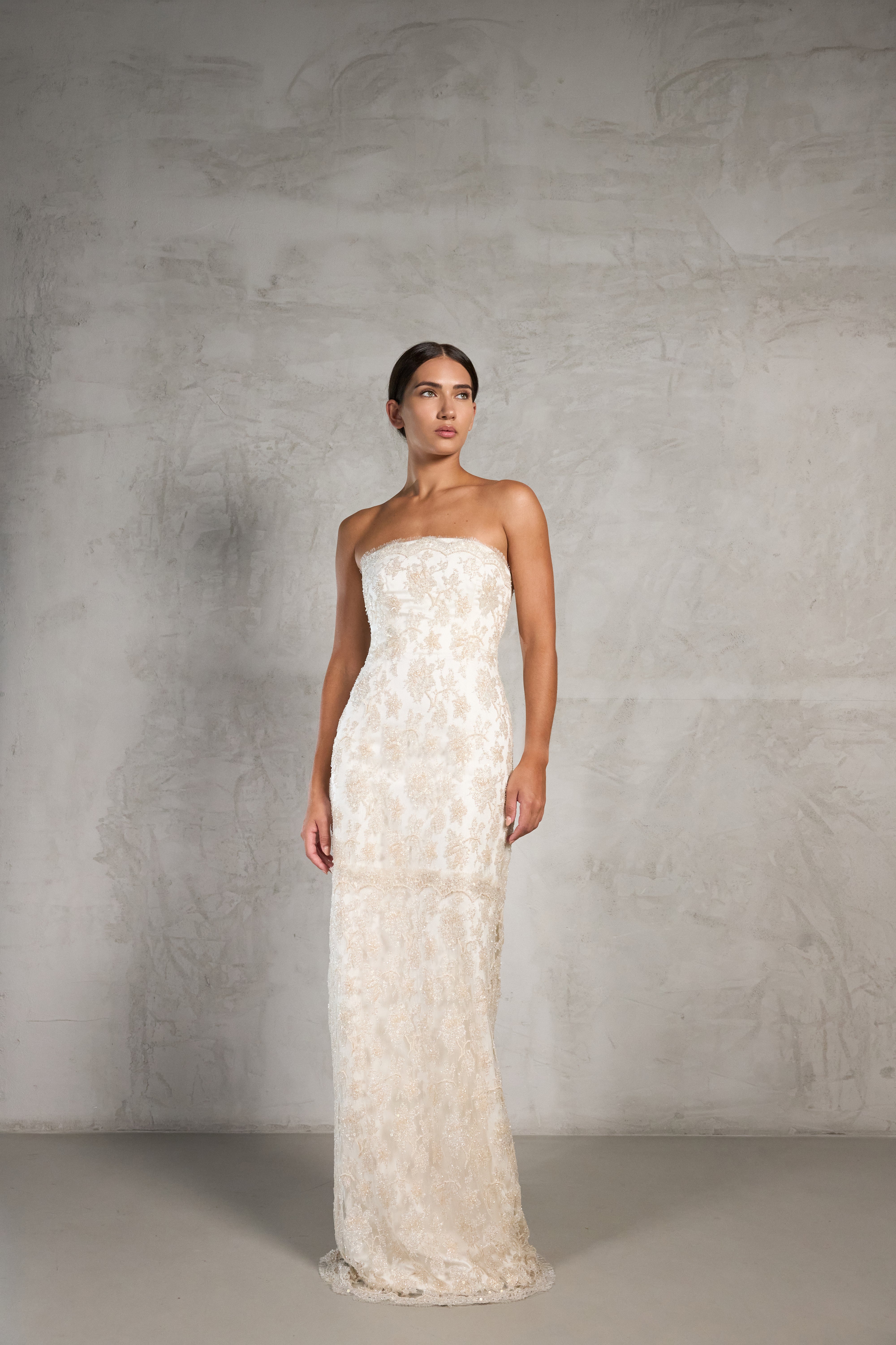 A woman modeling a bridal strapless, floor-length ivory lace gown against a textured gray studio backdrop.