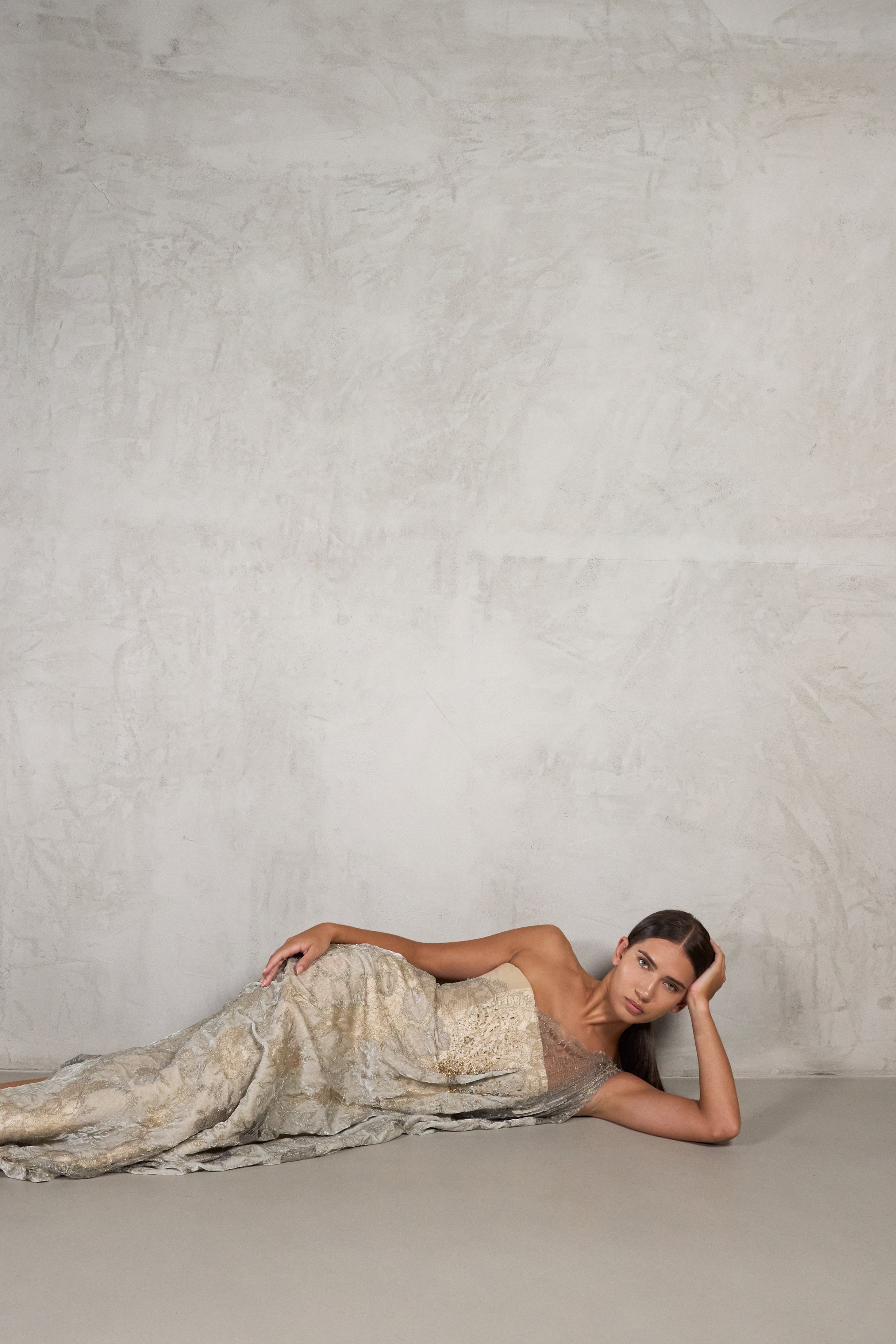 A woman reclines on a textured concrete floor against a matching concrete wall, wearing a strapless shimmering champagne-gold gown with a heavily embellished pattern.