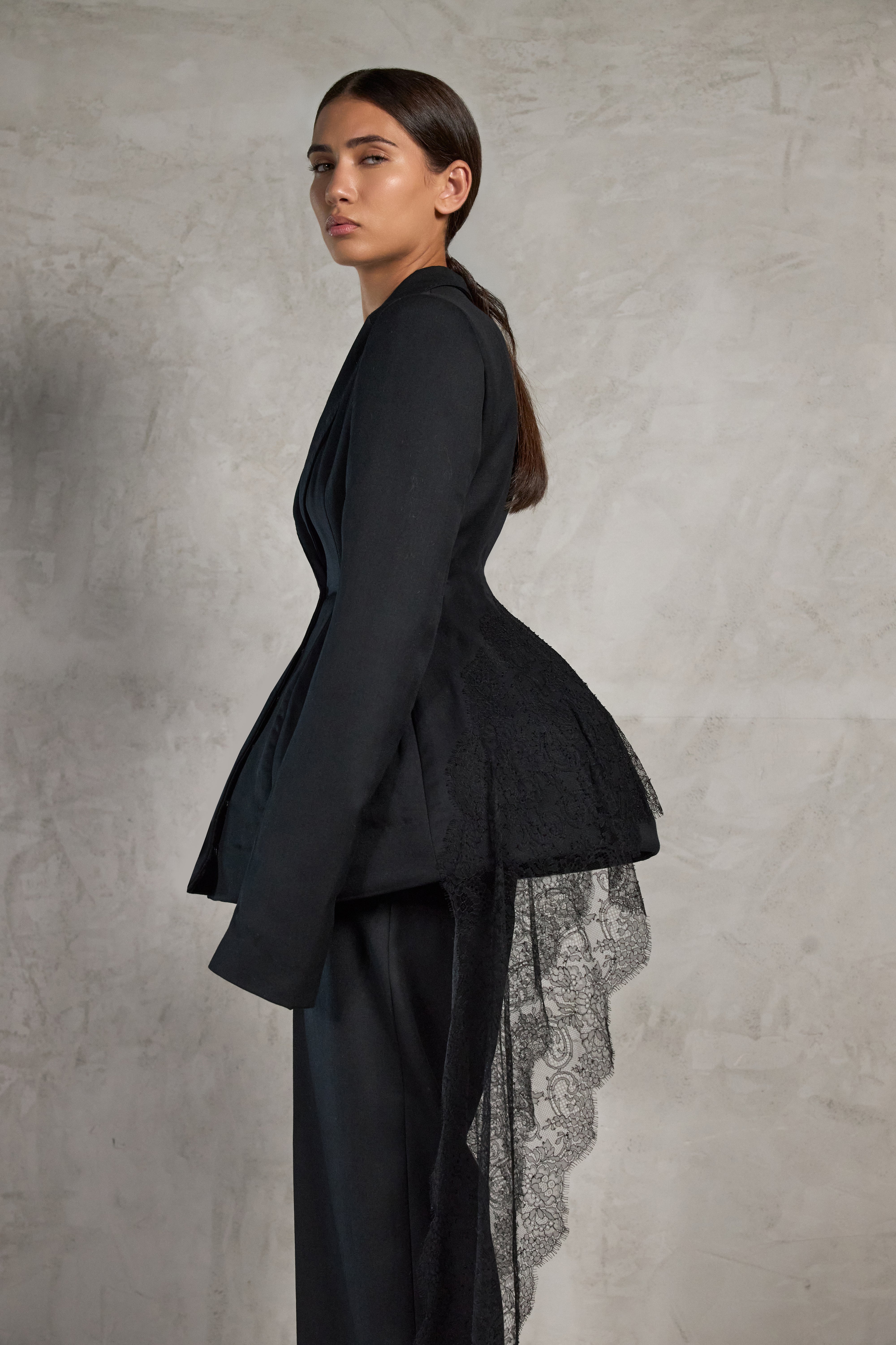 A model in side profile wearing a black structured jacket with a dramatic peplum waist, layered over a sheer black lace underlayer, inspired by the Maltese Ghonnella or Faldetta, set against a textured gray studio backdrop.
