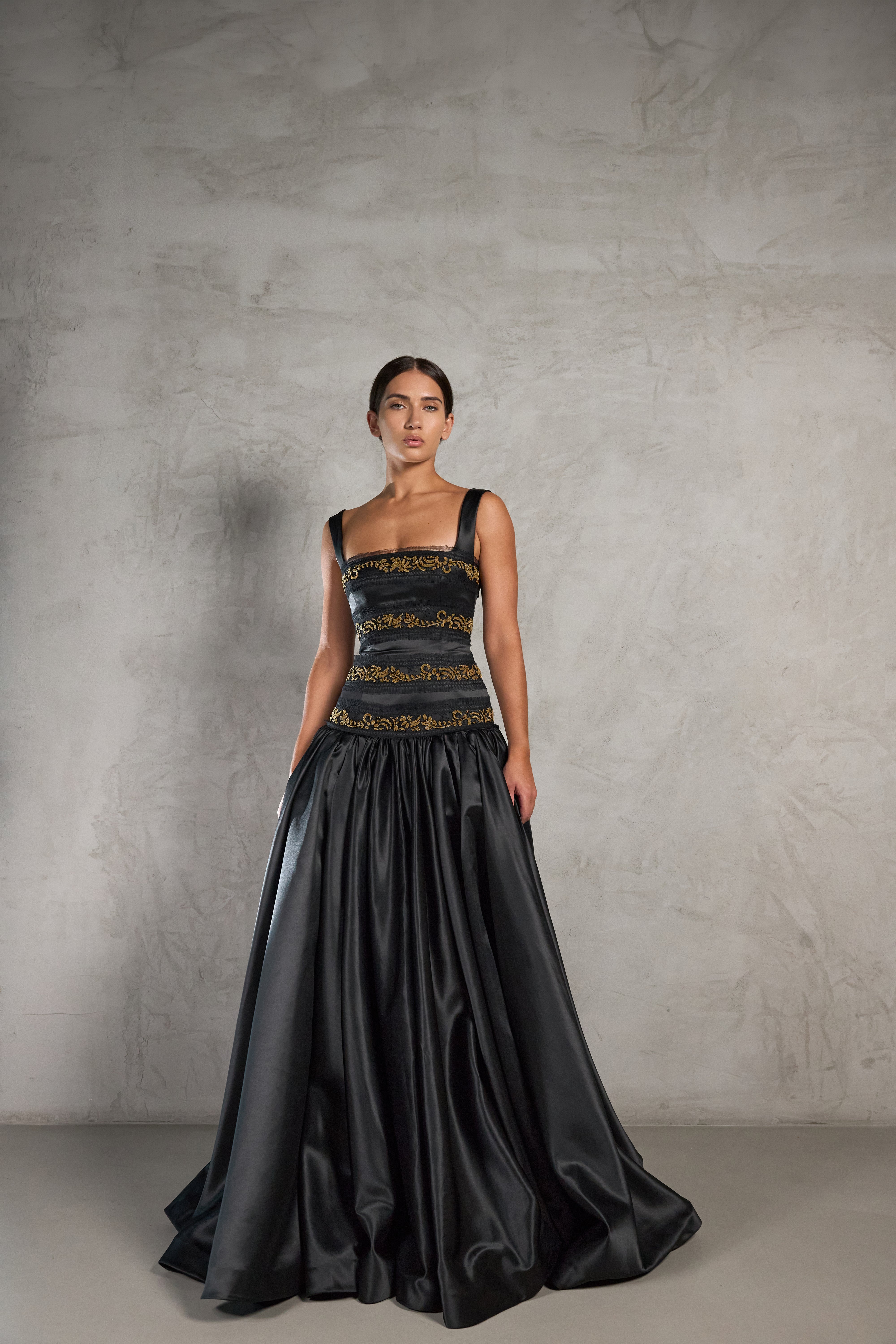 A model stands against a textured gray studio wall wearing a floor-length black gown with a structured bodice, gold embroidered horizontal bands, and a full satin-like skirt. The dress is Renaissance style. 
