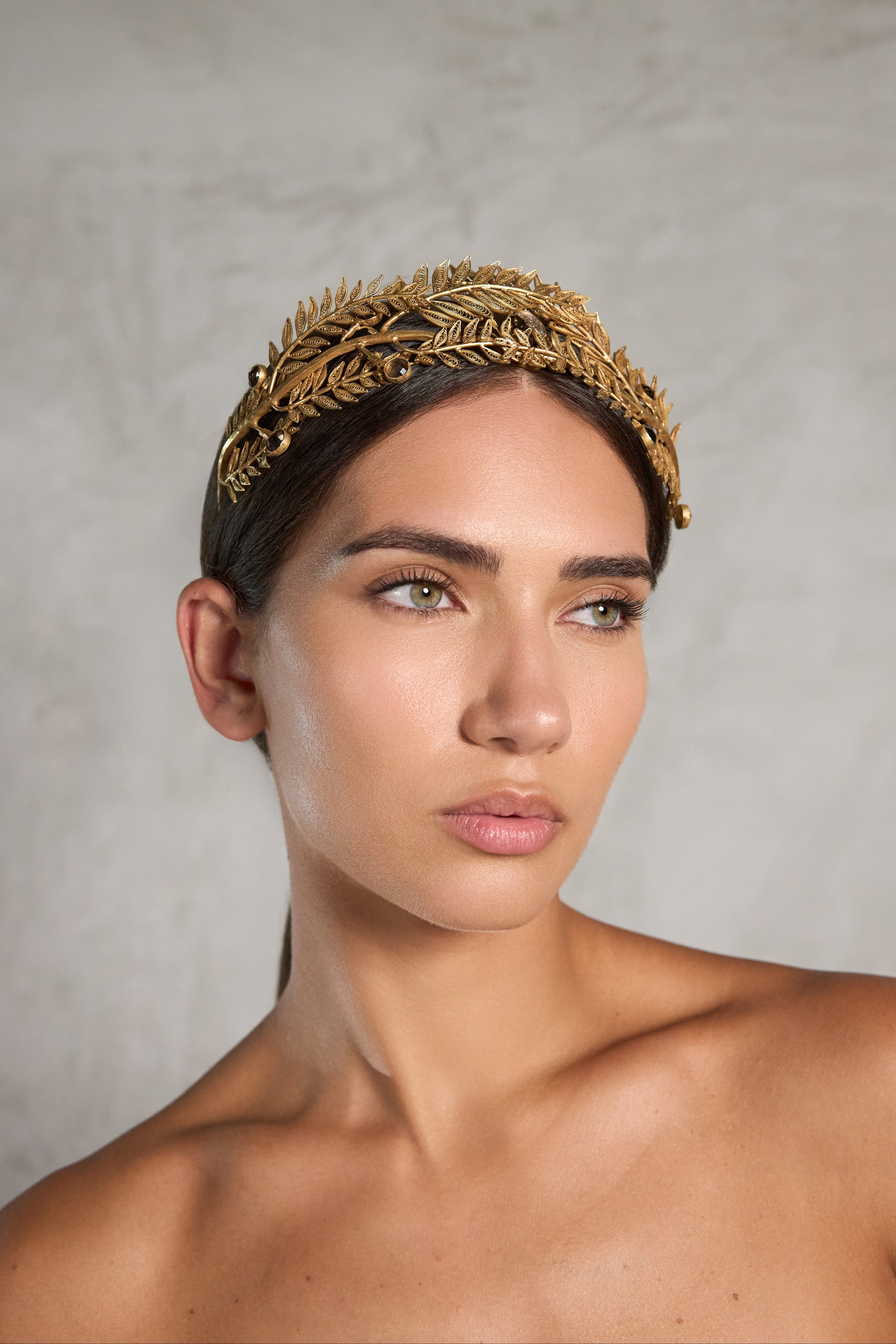A woman wearing an ornate gold leaf crown, photographed from the chest up against a textured light gray background.